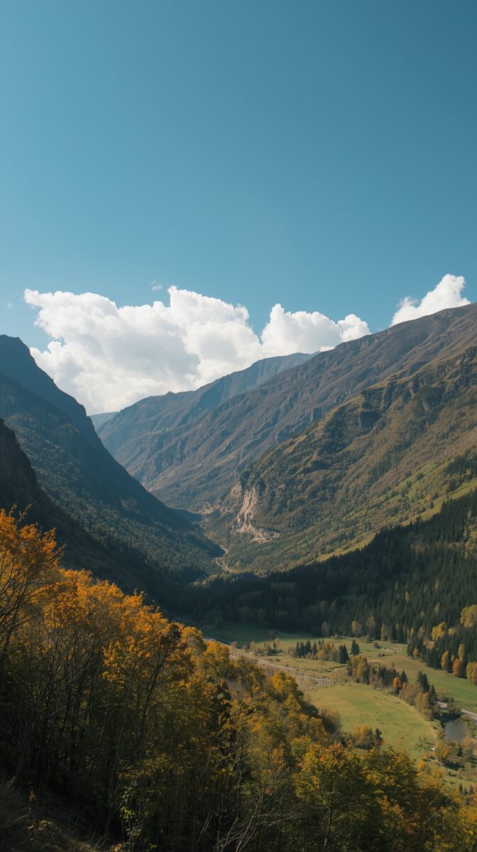Mountain valley in autumn with golden trees in foreground, dramatic peaks, and green meadow below under blue sky with clouds