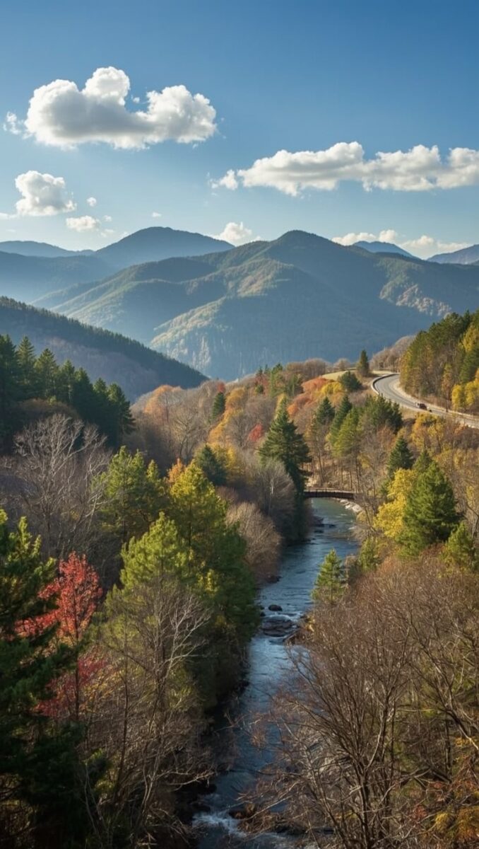Aerial view of a mountain valley with a winding river, autumn forest, curving highway, and blue ridge mountains under a partly cloudy sky
