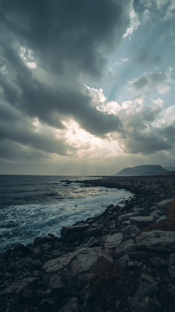 Dramatic rocky coastline with stormy skies, sunbeams breaking through clouds over crashing ocean waves and distant mountain
