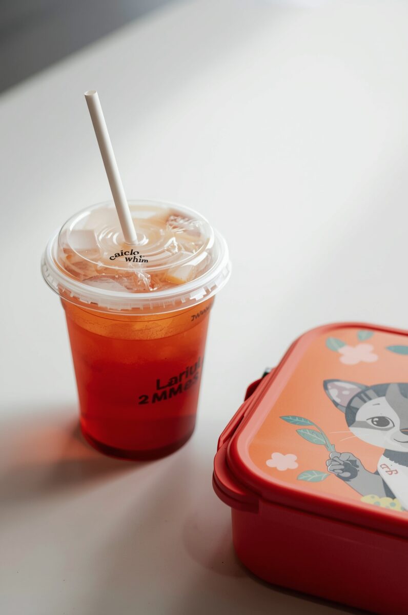 Iced fruit tea in clear plastic cup with white straw next to a red cartoon cat lunch box on white surface