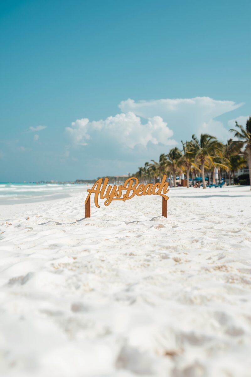 Wooden Alys Beach sign planted in white sand with palm trees and turquoise Gulf water in background