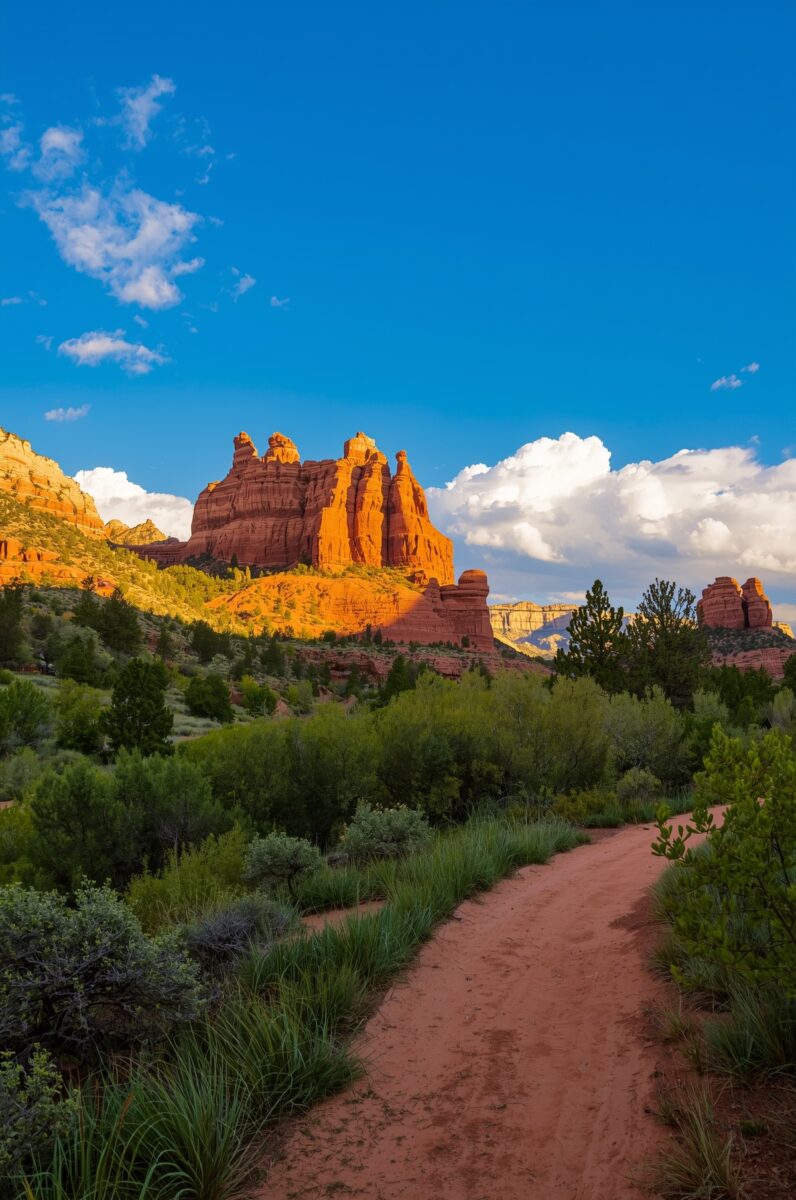 Red dirt trail winding through green desert shrubs toward towering orange-red sandstone buttes under blue sky