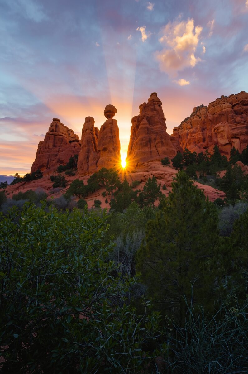 Sunburst between red sandstone rock spires at sunset with green trees in foreground, American Southwest