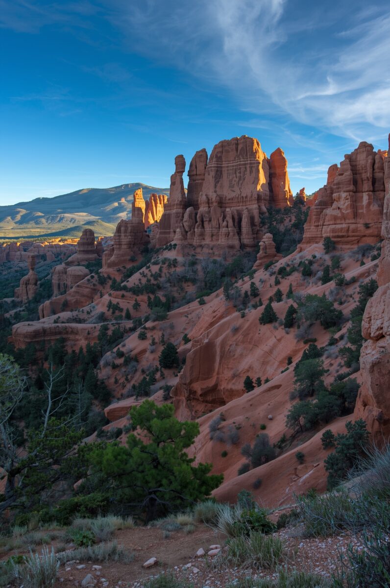 Red sandstone spires and hoodoos rise above a canyon landscape with scattered pine trees under a blue sky in Utah