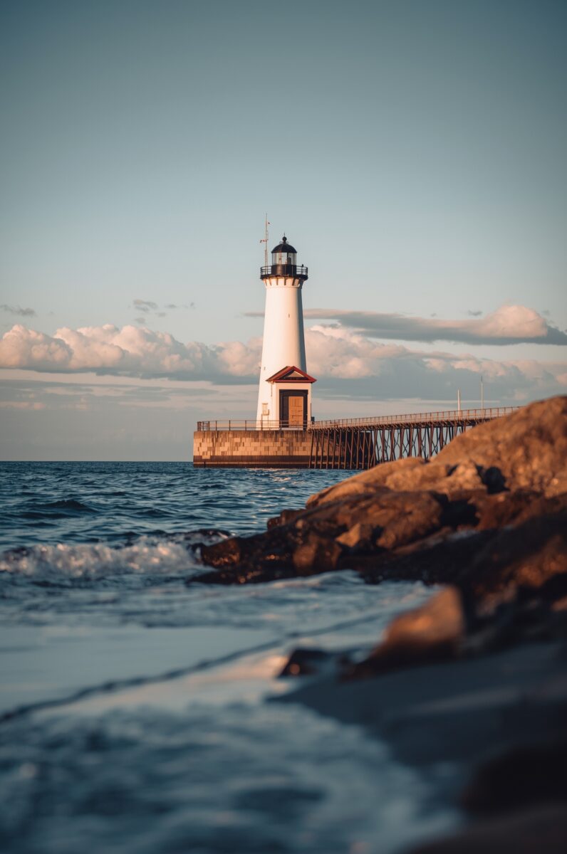 White lighthouse on a stone pier extending into calm blue waters under a partly cloudy sky at golden hour