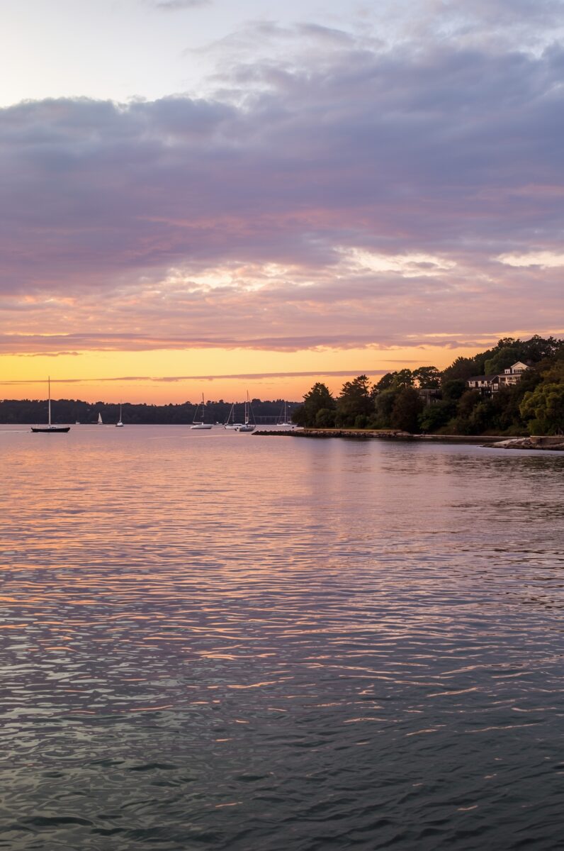 Sailboats moored in a calm harbor at sunset with purple and orange sky reflecting on water