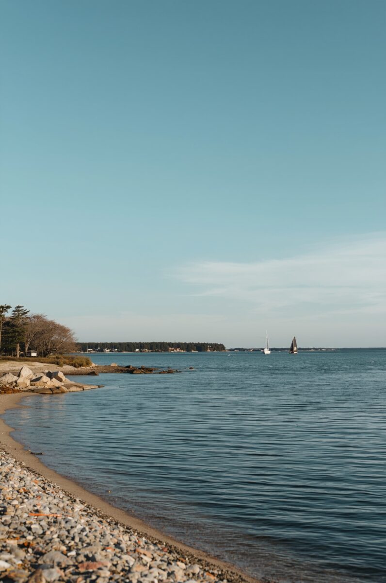 Rocky shoreline with calm blue water and two sailboats in the distance under a clear sky