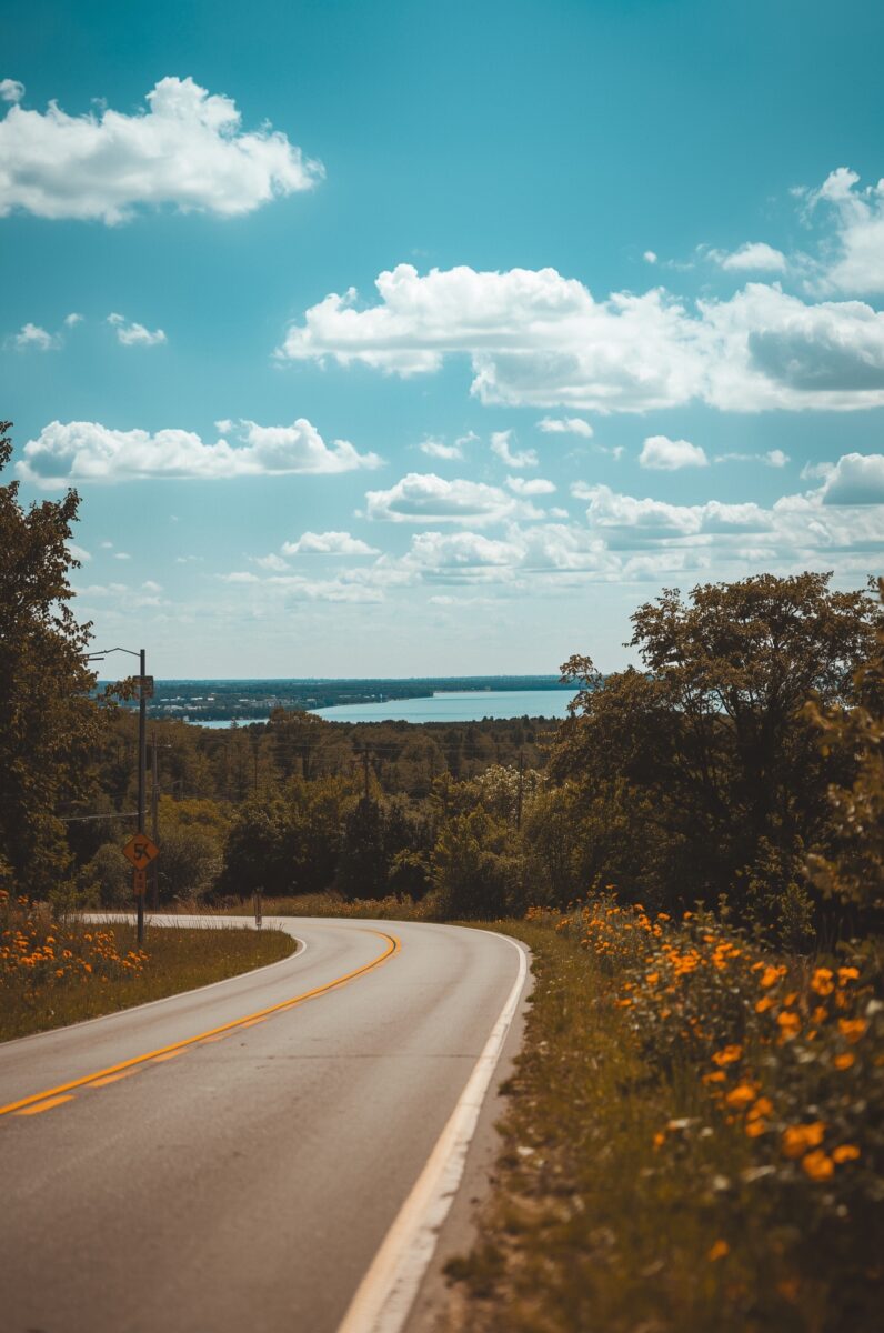 Winding two-lane rural road curving downhill through lush green trees toward a distant lake under blue skies