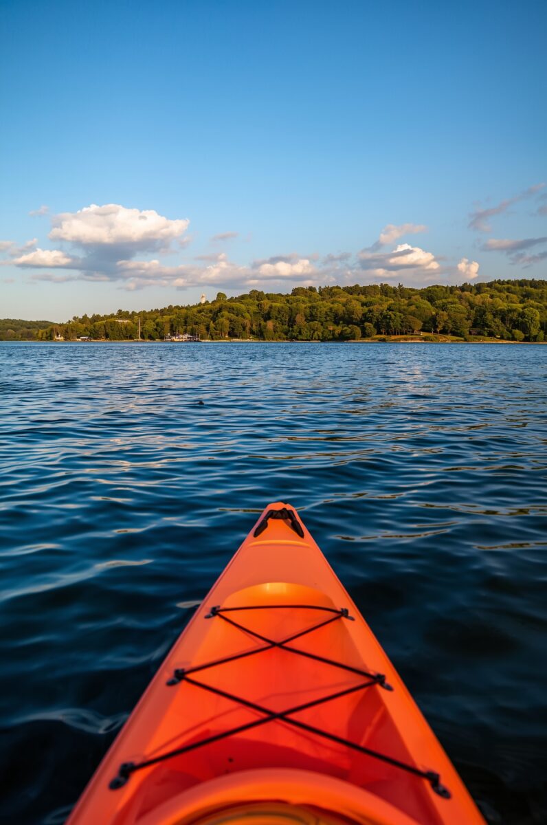 Orange kayak bow pointing toward a tree-lined shoreline on a calm blue lake under a partly cloudy sky