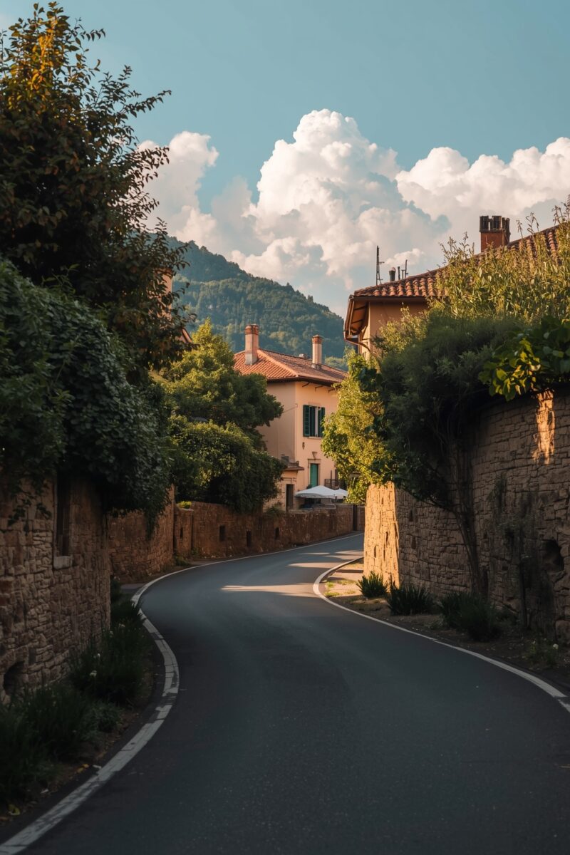 Winding narrow road flanked by stone walls and lush greenery leading to Italian village buildings with terracotta roofs and mountain backdrop