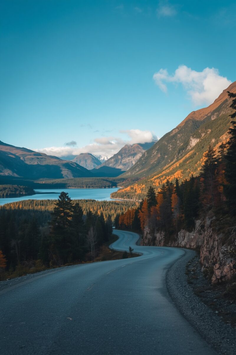 Winding mountain road curving through autumn forest with turquoise lake and snow-capped peaks in background