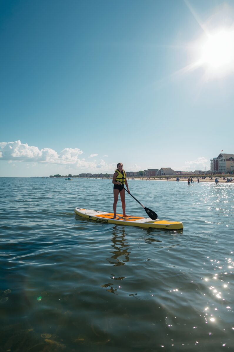 Young woman in yellow life jacket standing on yellow paddleboard on calm ocean water near a beach town