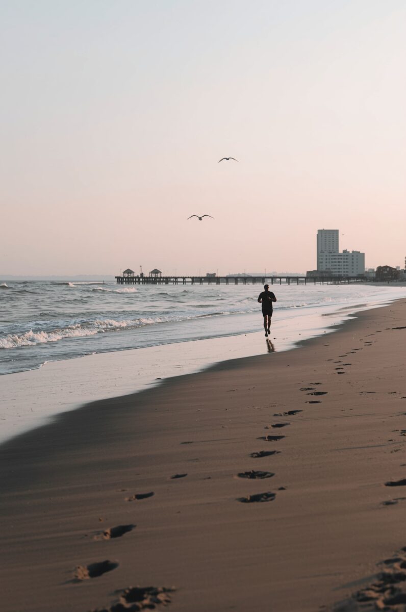 Lone runner jogging along a sandy beach at sunrise with a pier and city buildings in the background