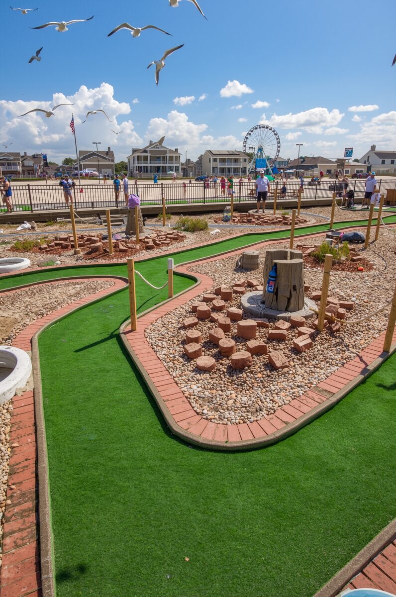Colorful mini golf course with log stump obstacles, gravel decorations, and a Ferris wheel visible in the background near a beachside boardwalk