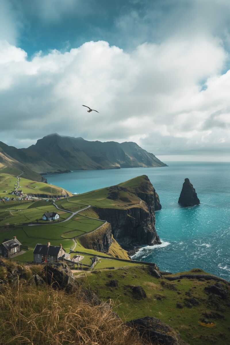 Dramatic coastal cliffside with green fields, stone cottages, a sea stack, and turquoise ocean under a cloudy sky