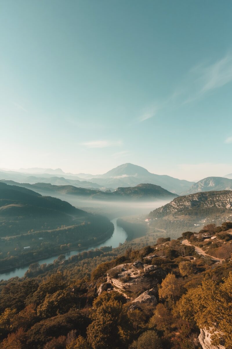 Aerial view of a winding river valley surrounded by misty mountains and lush forested hills under a clear blue sky