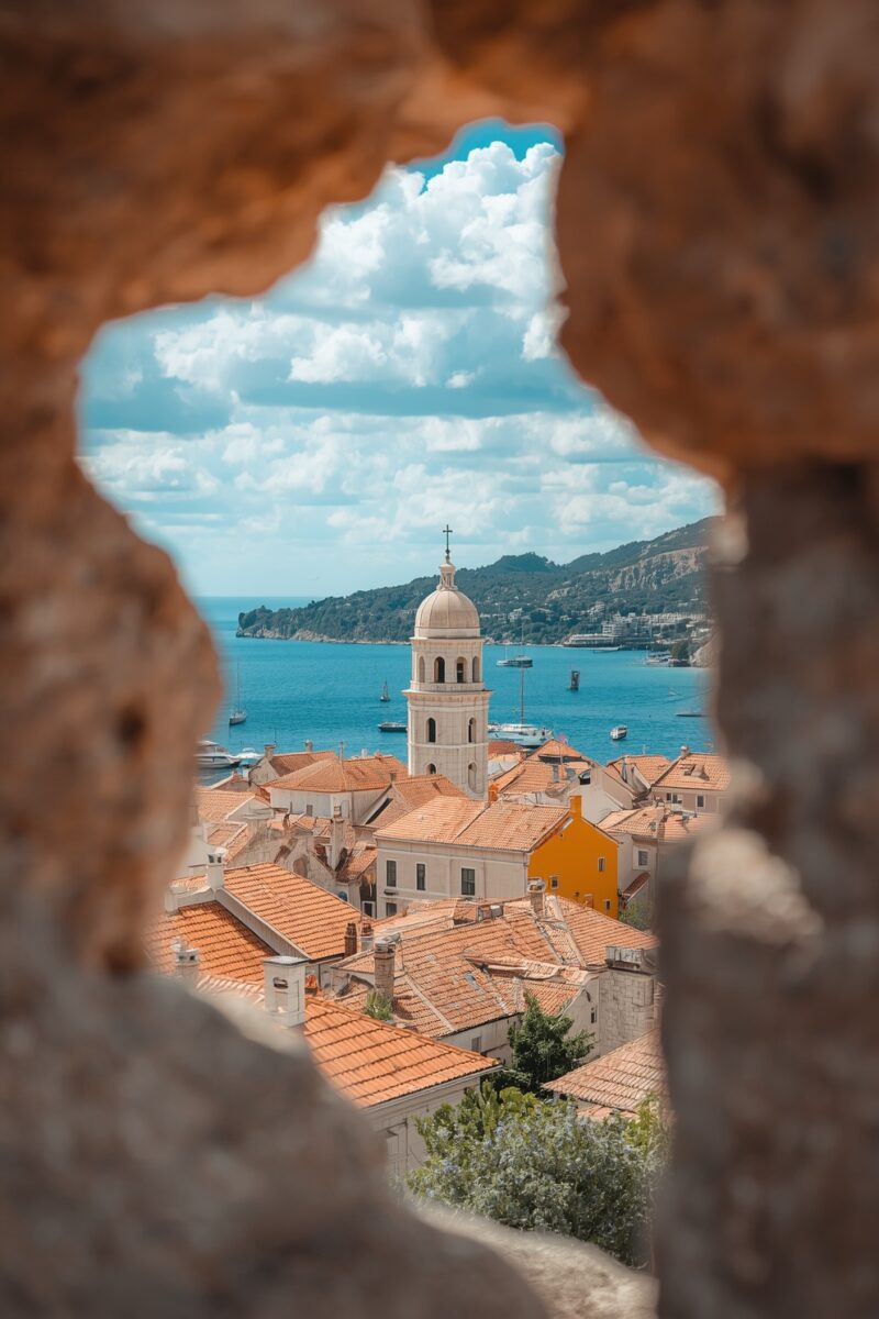 View through stone wall opening of Mediterranean coastal town with terracotta rooftops, church bell tower, and blue Adriatic Sea
