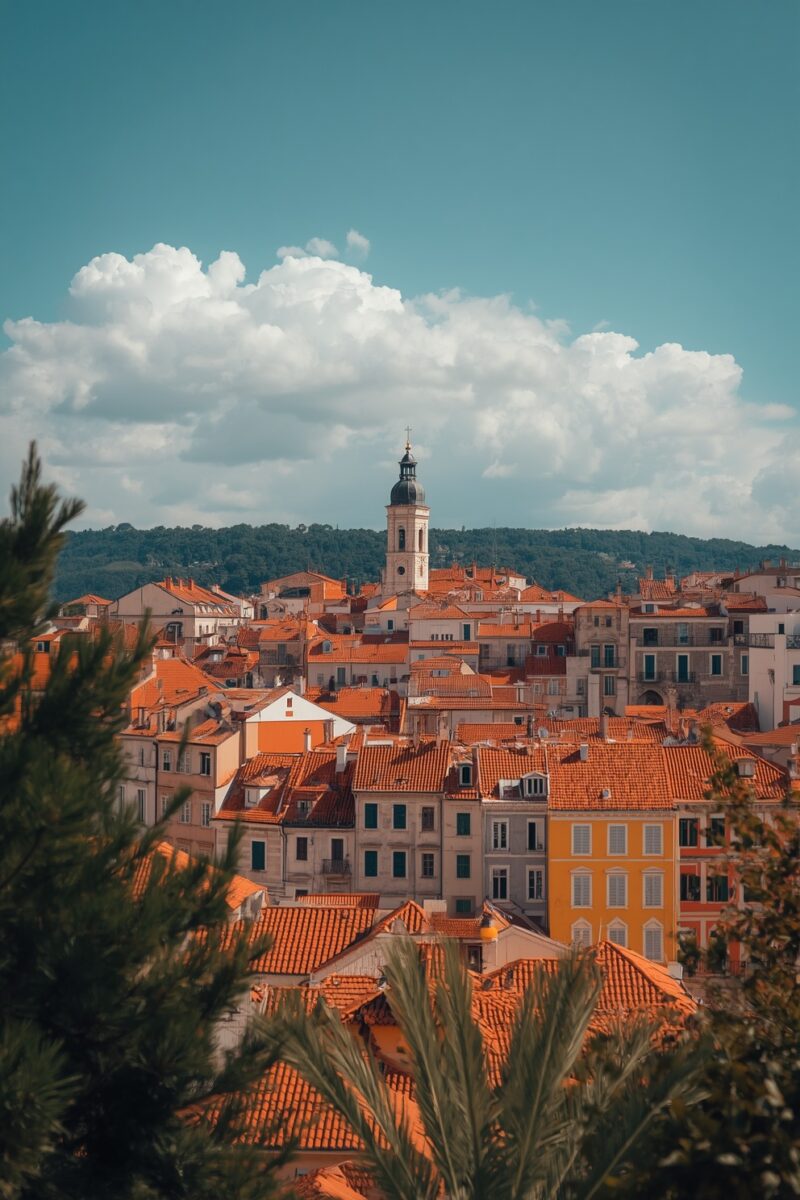 Panoramic view of a Mediterranean old town with orange terracotta rooftops, a white church bell tower, and green hills behind