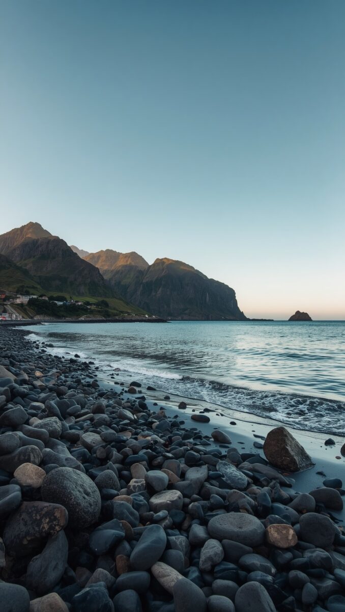 Rocky pebble beach with calm sea, dramatic mountains, and clear blue sky at dusk in Norway