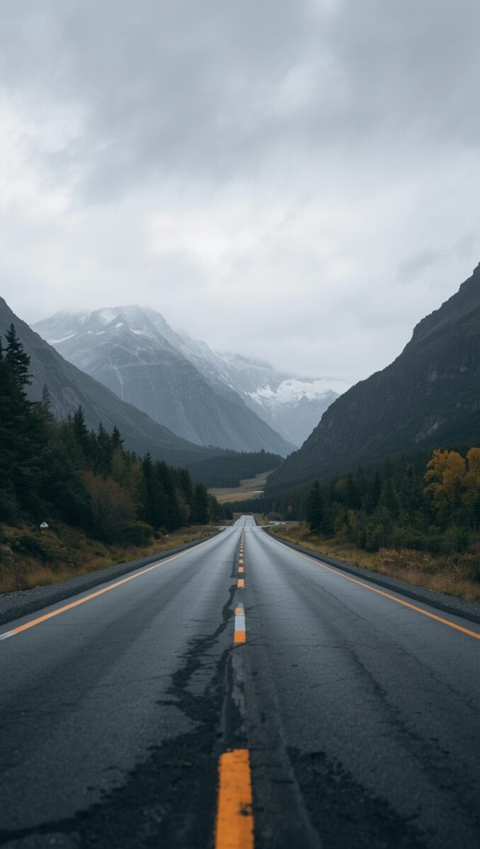 Empty two-lane highway stretching into a misty mountain valley with snow-capped peaks and autumn trees