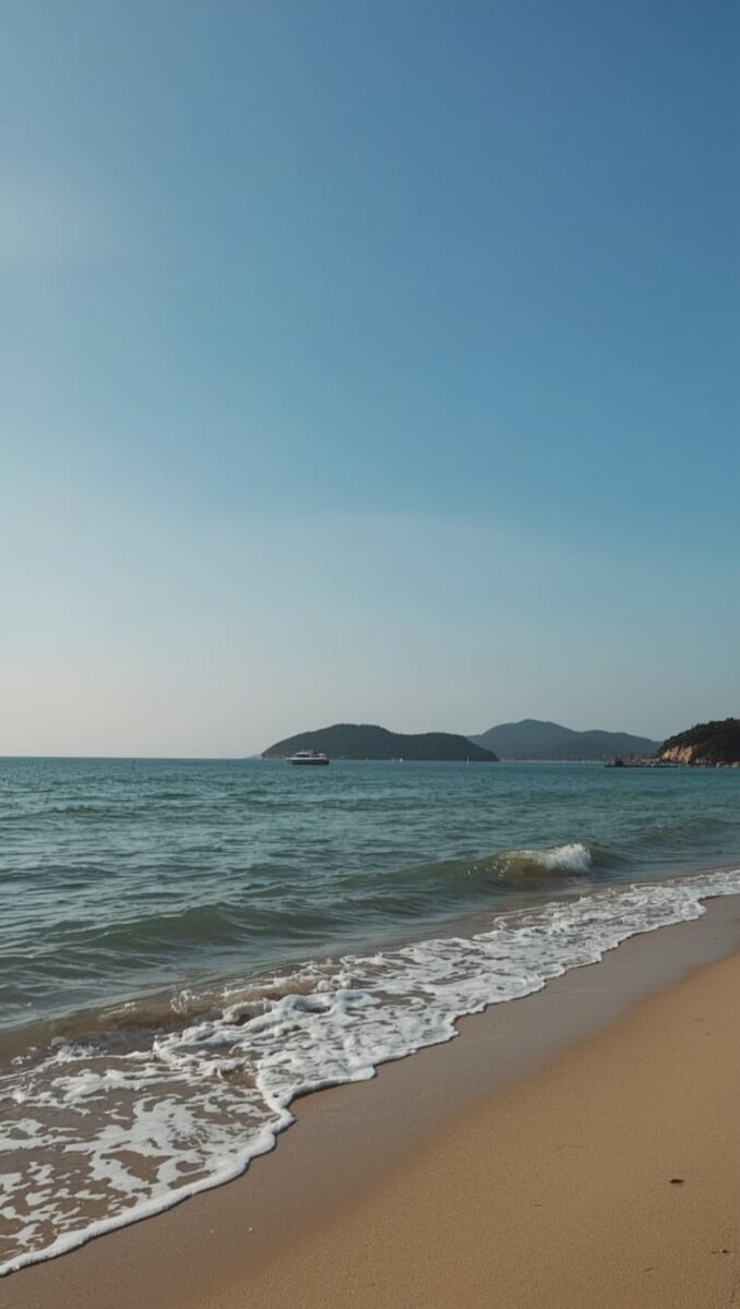 Sandy beach with gentle waves, turquoise sea, distant green islands, and a clear blue sky