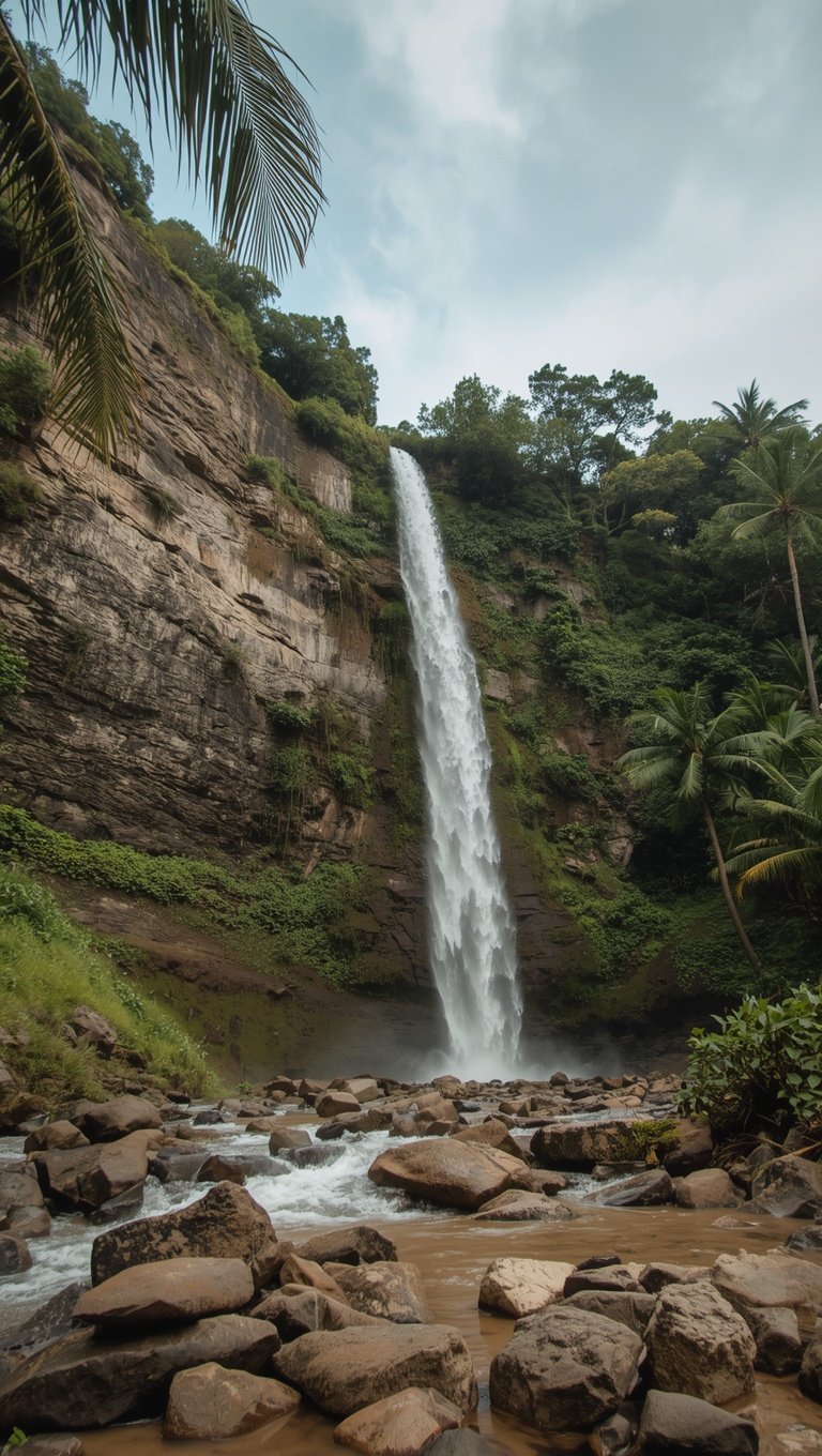 Tall single-drop waterfall cascading down a rocky cliff face surrounded by tropical jungle and palm trees