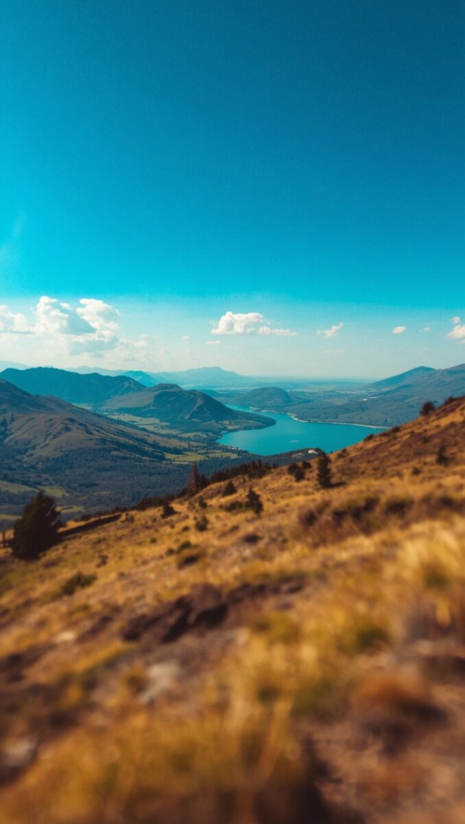 Panoramic mountain landscape with turquoise lake in valley, golden hillside foreground, clear blue sky