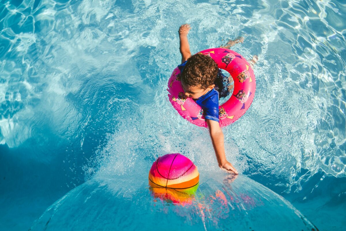 A small child in a pink swim ring reaching for a bright beach ball in a pool