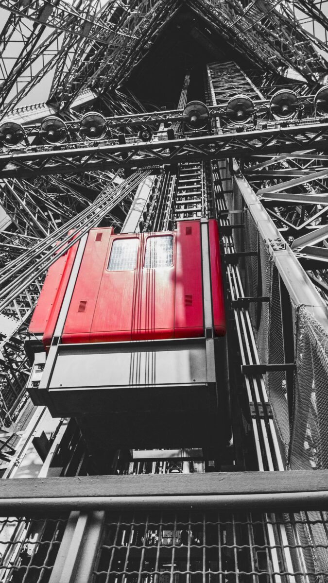 A red Eiffel Tower lift car on its rails inside the iron framework, in a black and white photo with only the car in color