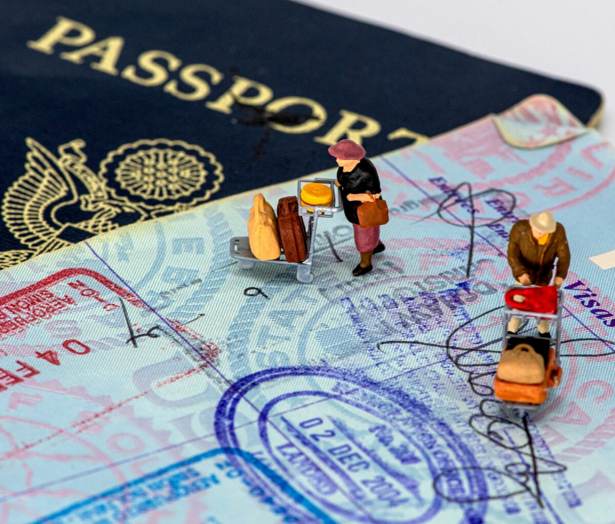 Two tiny travelers with luggage carts standing on an open US passport full of entry stamps