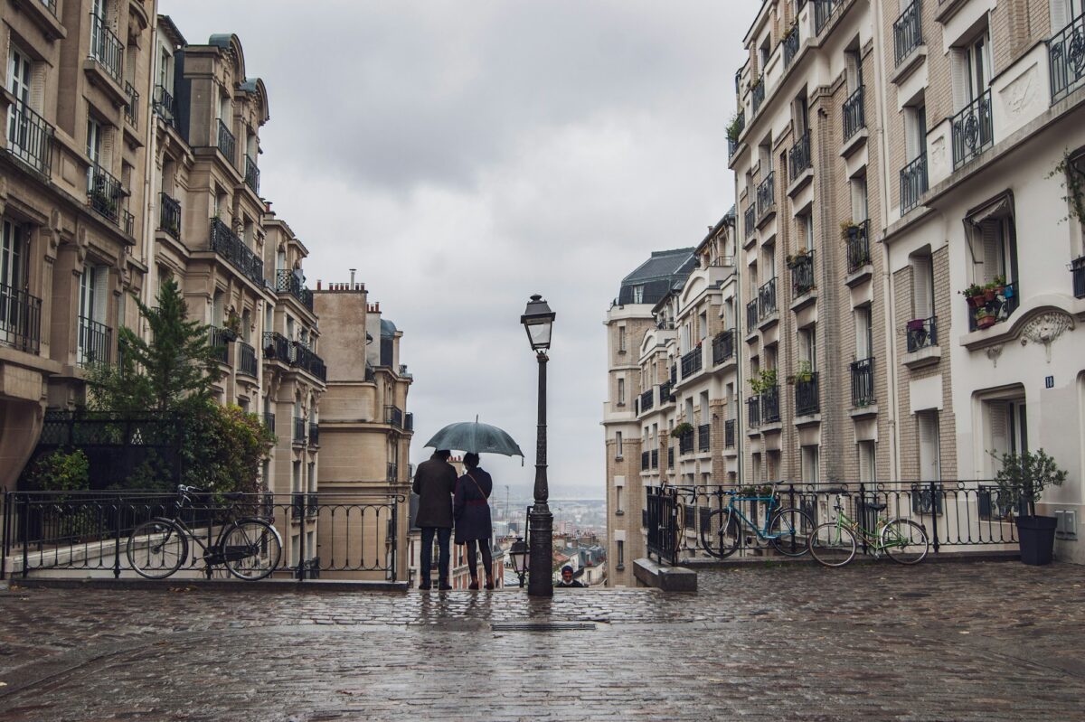 Two people under an umbrella on a wet cobbled square in Paris, looking out over the city on a gray day