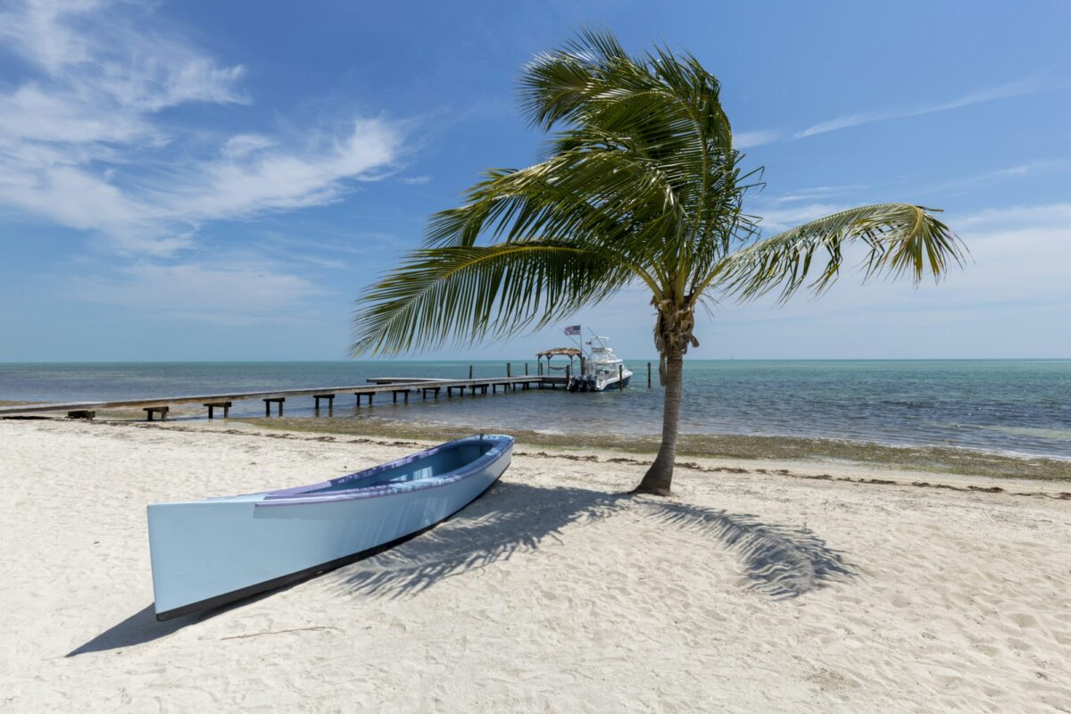 A palm tree and a pale blue canoe on white sand, with a fishing boat at a long dock in Islamorada, Florida Keys