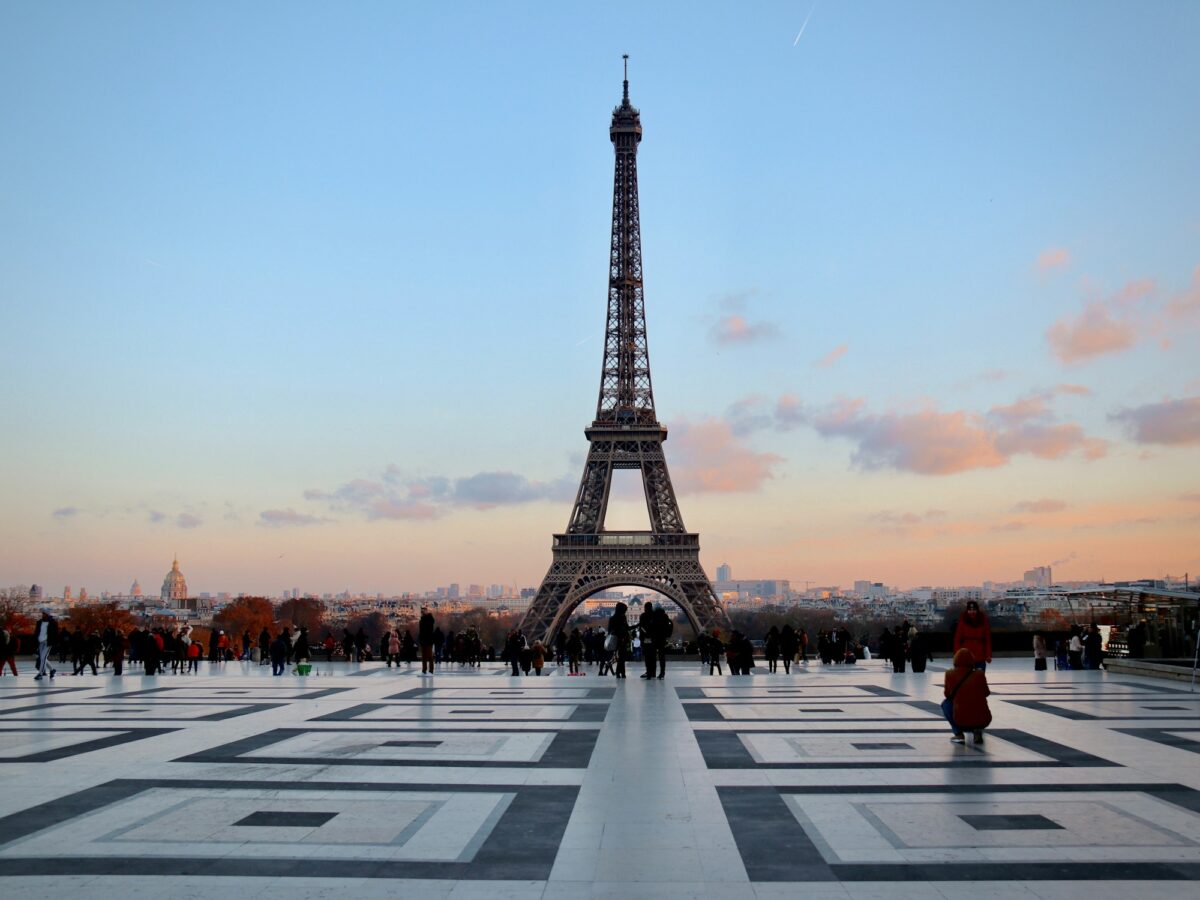 People in winter coats on the Trocadero esplanade at dusk, with the Eiffel Tower against a pink sky