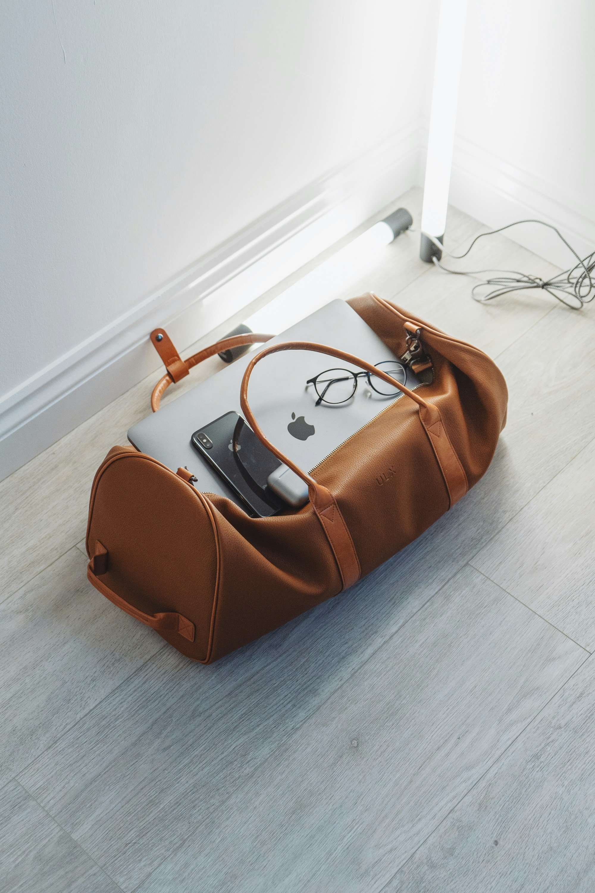 An open tan leather duffel on a gray wood floor holding a silver MacBook, an iPhone plugged into a small white power bank, round glasses and a cable