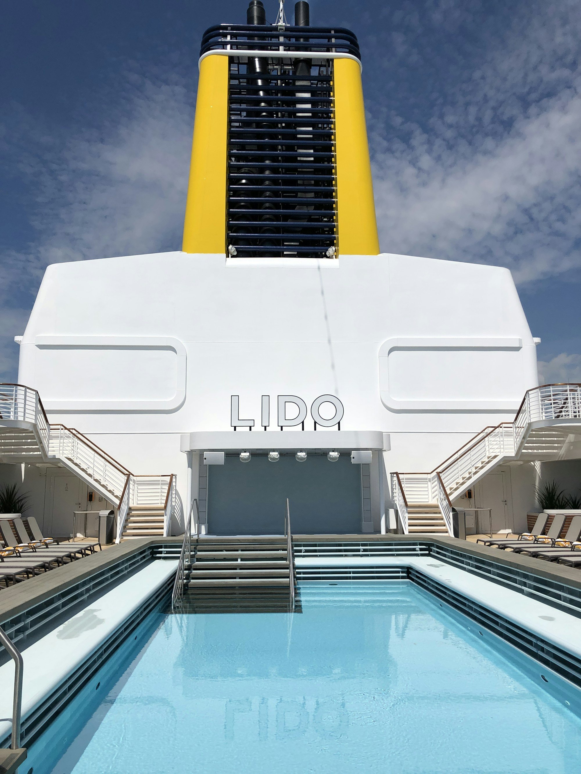 A symmetrical LIDO pool on a cruise ship with the LIDO sign reflected in the water, a yellow funnel and blue sky above