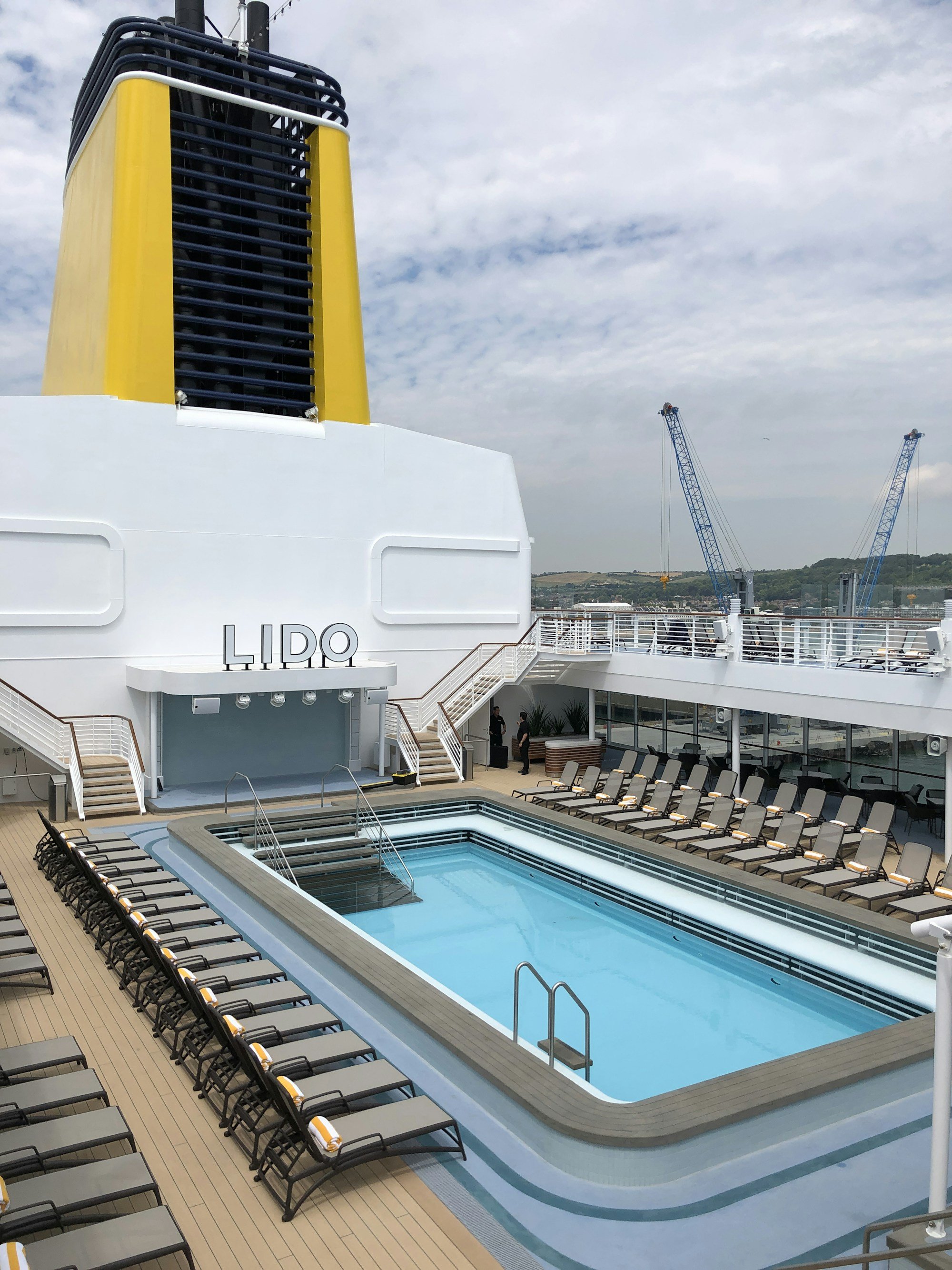 A cruise ship's LIDO pool deck with rows of empty loungers and a yellow funnel, port cranes visible behind