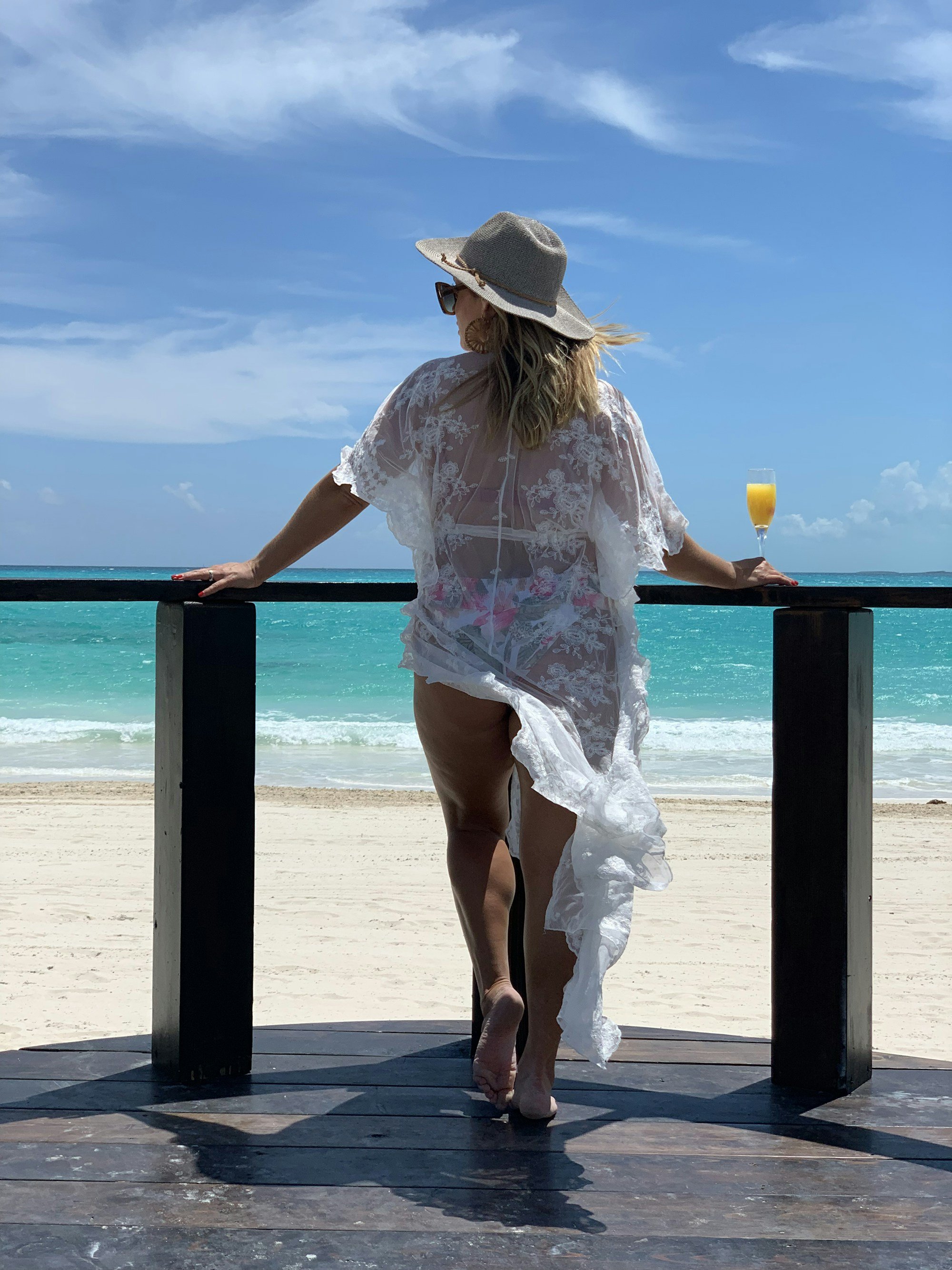 A woman seen from behind at a deck railing in a sheer white lace kimono cover-up over a swimsuit, wearing a fedora and holding an orange drink, turquoise sea below