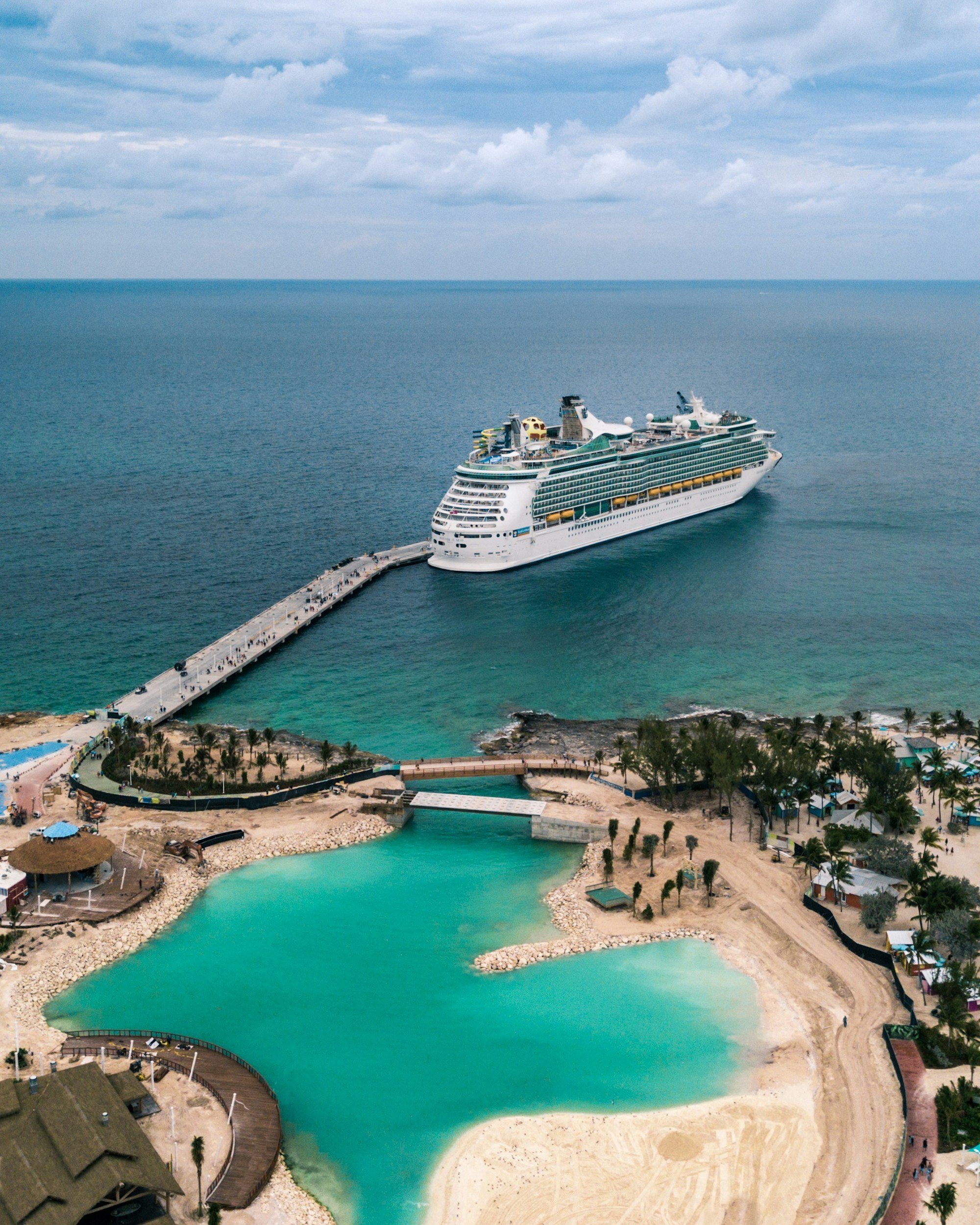 Aerial view of a cruise ship docked at a long pier beside a turquoise lagoon lined with palm trees and huts