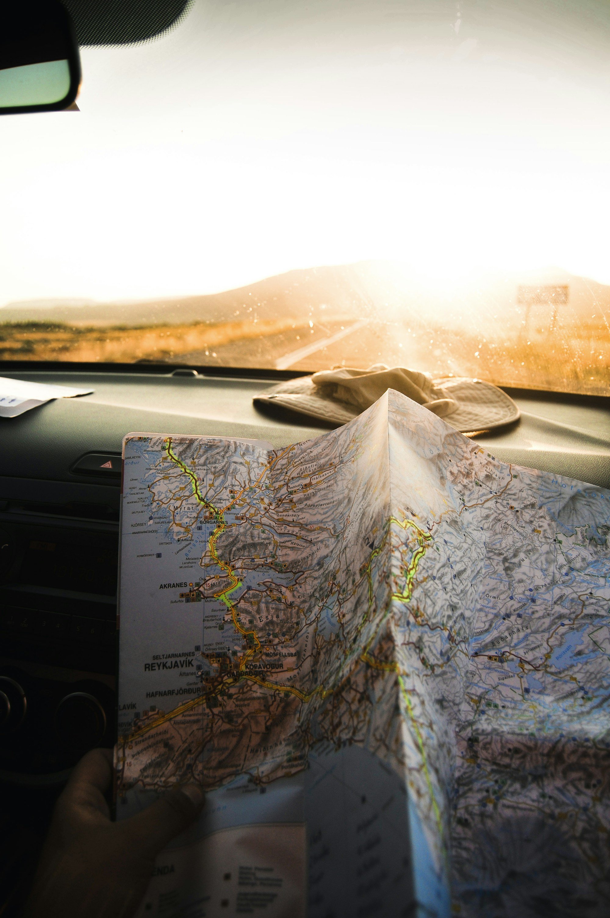 A paper map of Iceland held up against low sun glare through a car windshield, a bucket hat on the dashboard