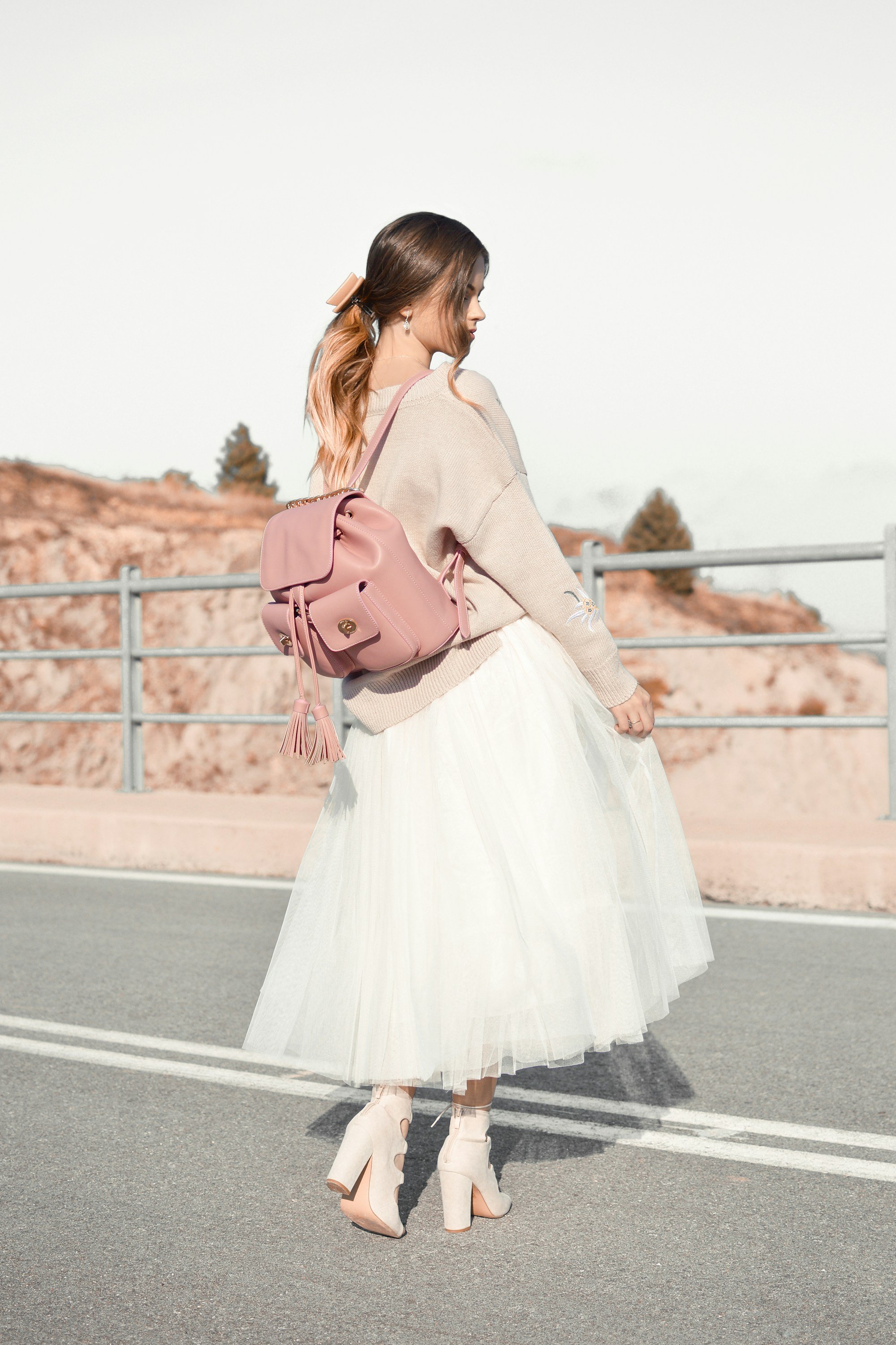 A woman in a beige sweater, a white tulle midi skirt and blush block-heel ankle boots with a small pink drawstring backpack, standing on a road by a guardrail