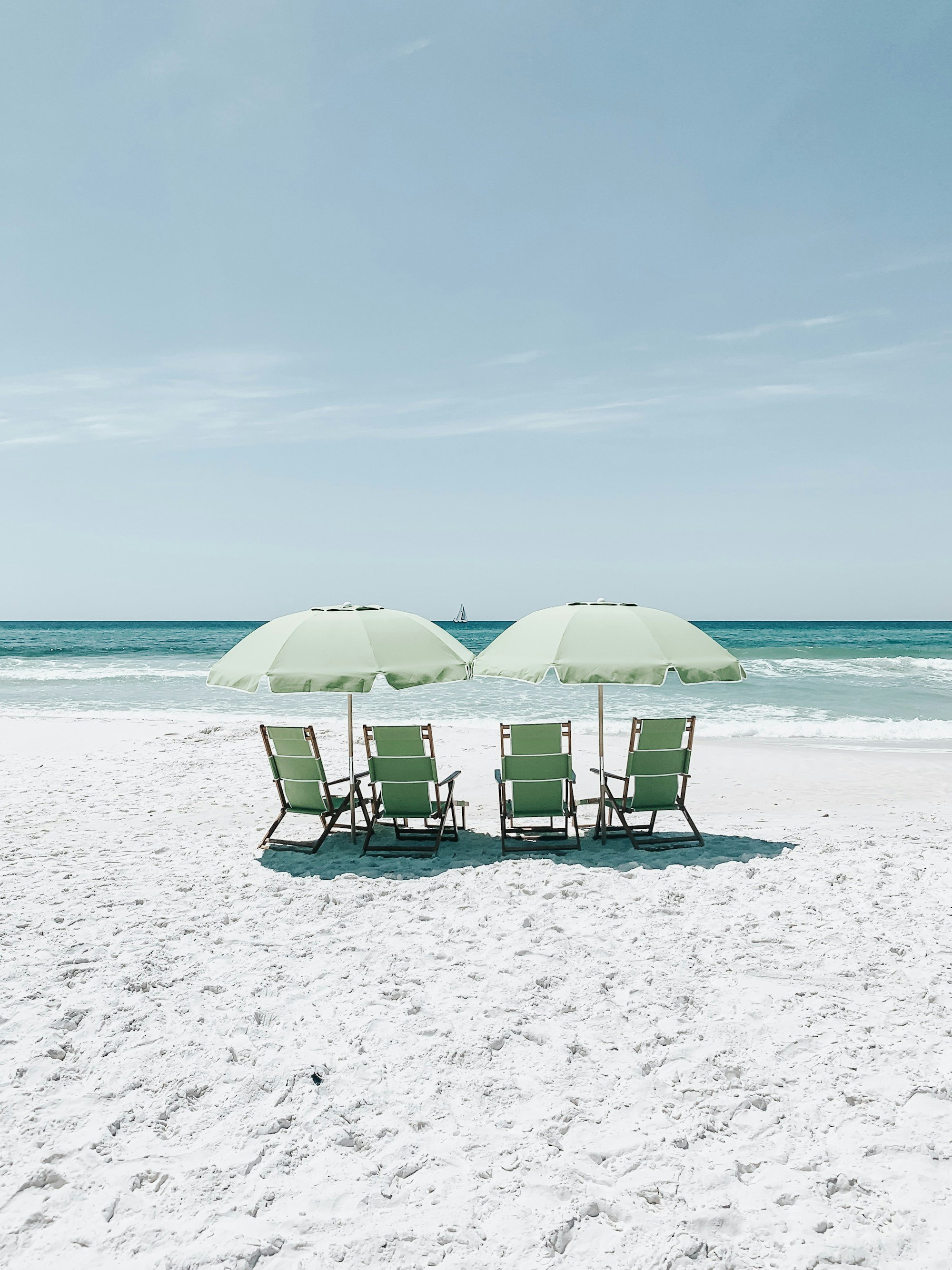 Two pale green beach umbrellas over four green lounge chairs on white sand, turquoise water and a sailboat behind