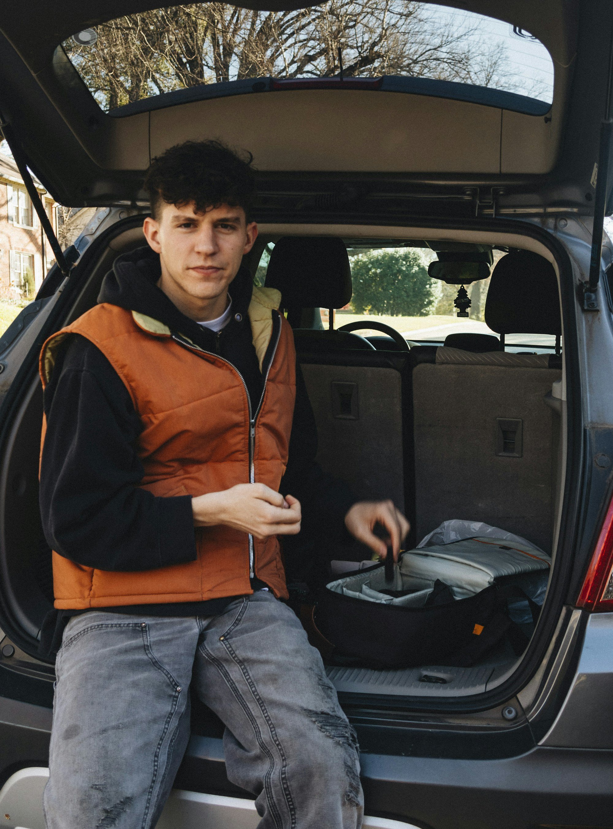 A young man in an orange puffer vest sits on the open back of a small SUV beside an open, padded soft bag