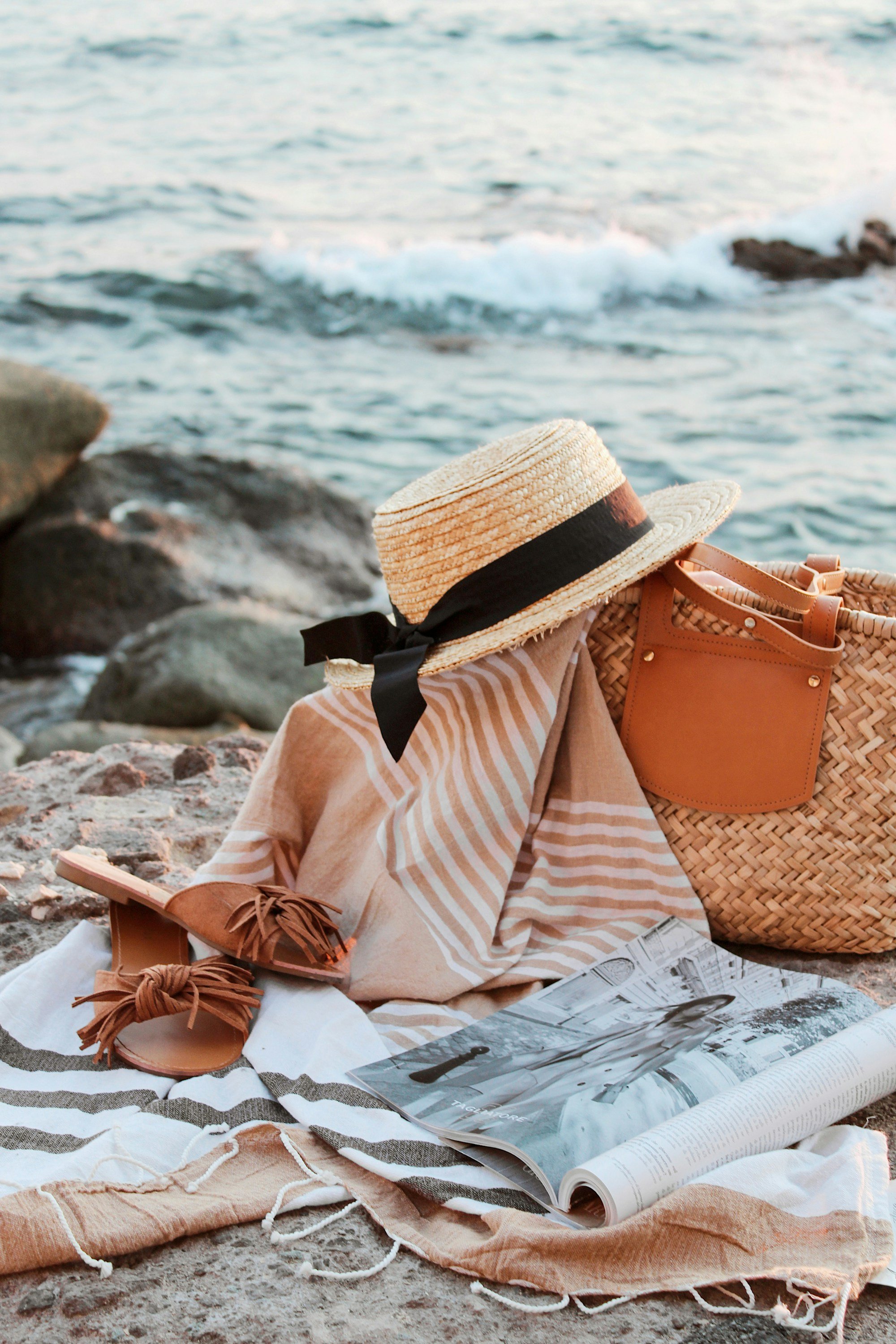 A thin striped towel with fringed ends draped under a straw boater hat, a straw basket and leather slides beside it, waves breaking on rocks behind