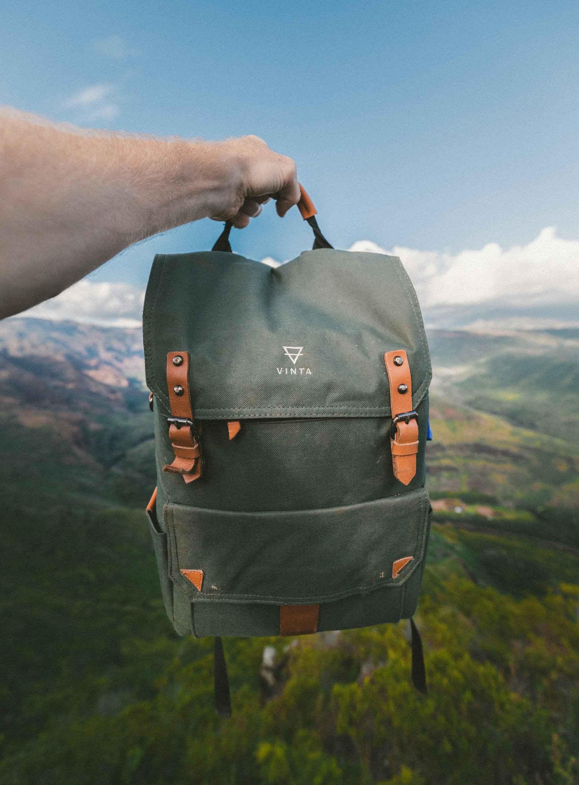 A hand holding up an olive canvas backpack with tan leather straps and buckles against a green mountain valley