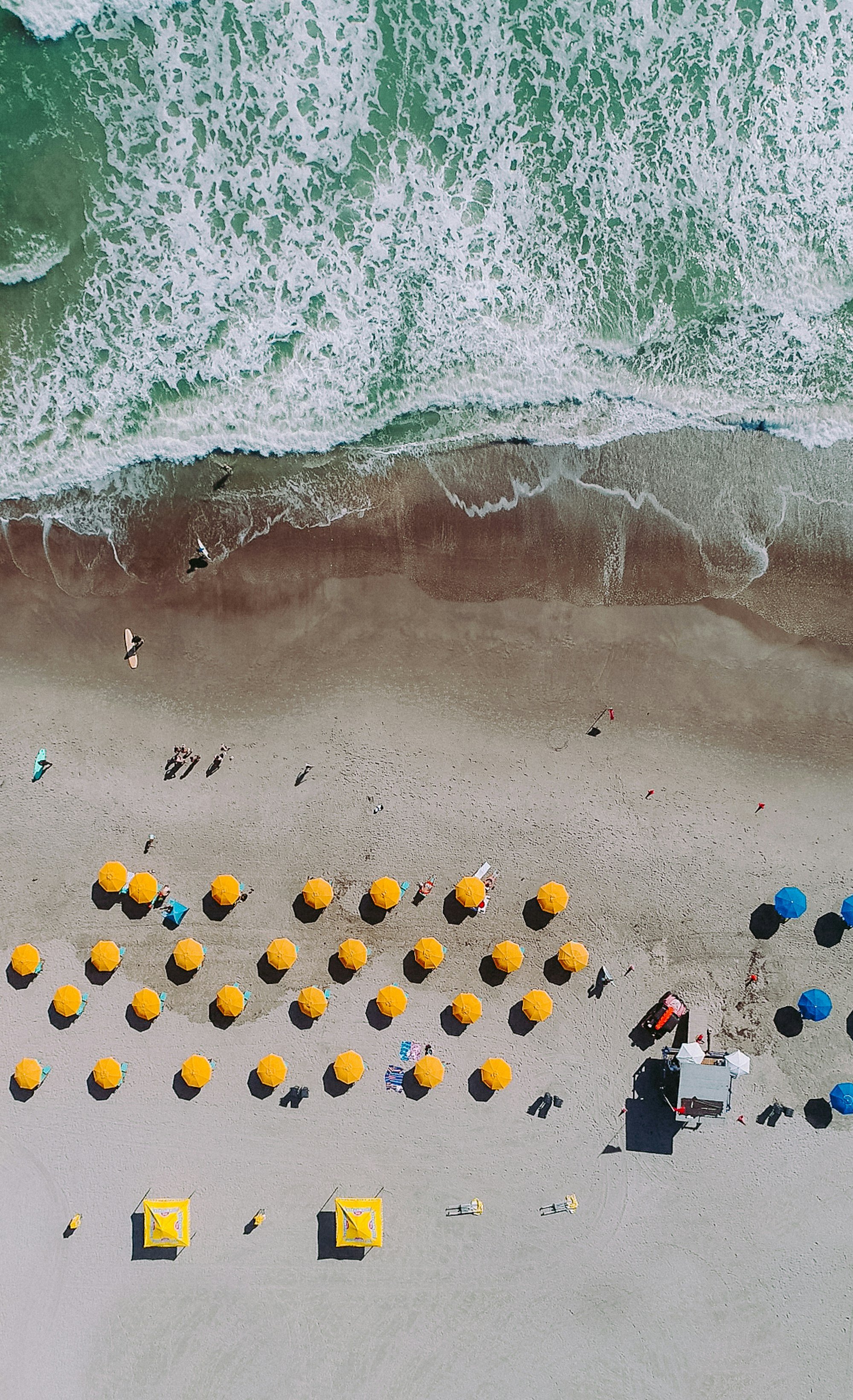 Aerial view of rows of yellow rental umbrellas and a lifeguard stand above the surf, people standing at the waterline