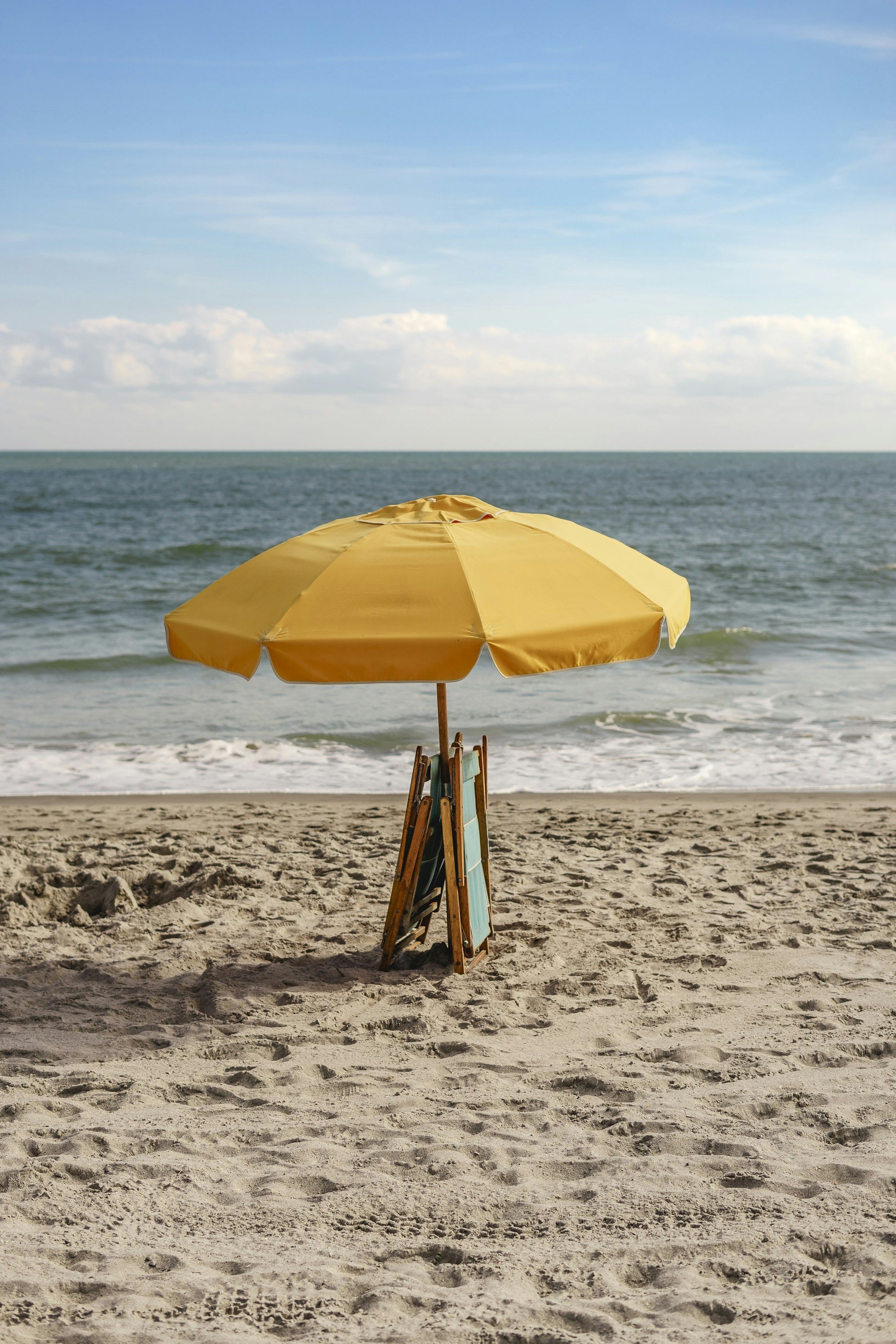 A single yellow beach umbrella on an empty beach with folding wooden chairs leaning against its pole