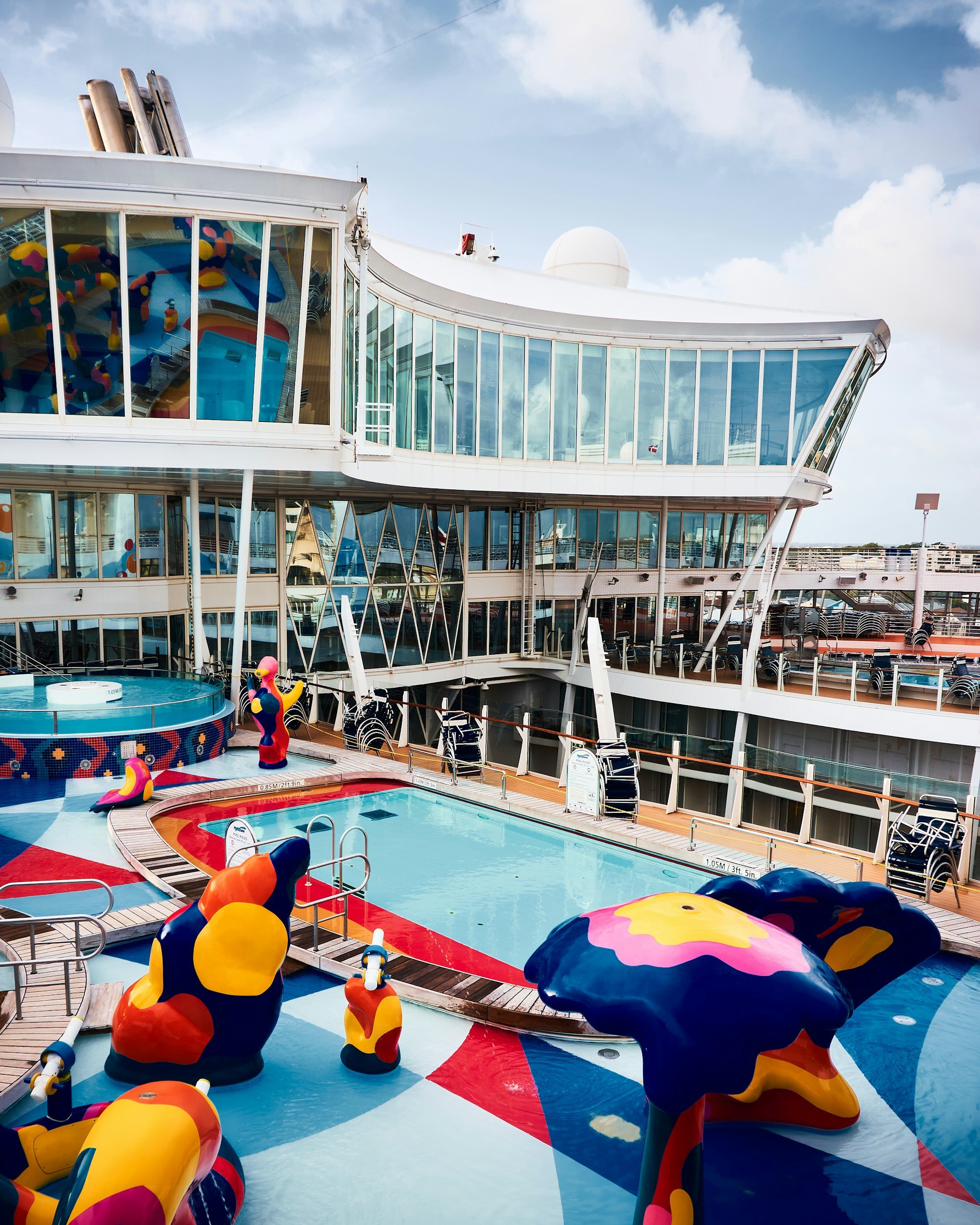 A colorful cruise ship pool deck with bright painted sculptures, a hot tub, stacked loungers and glass-sided decks above