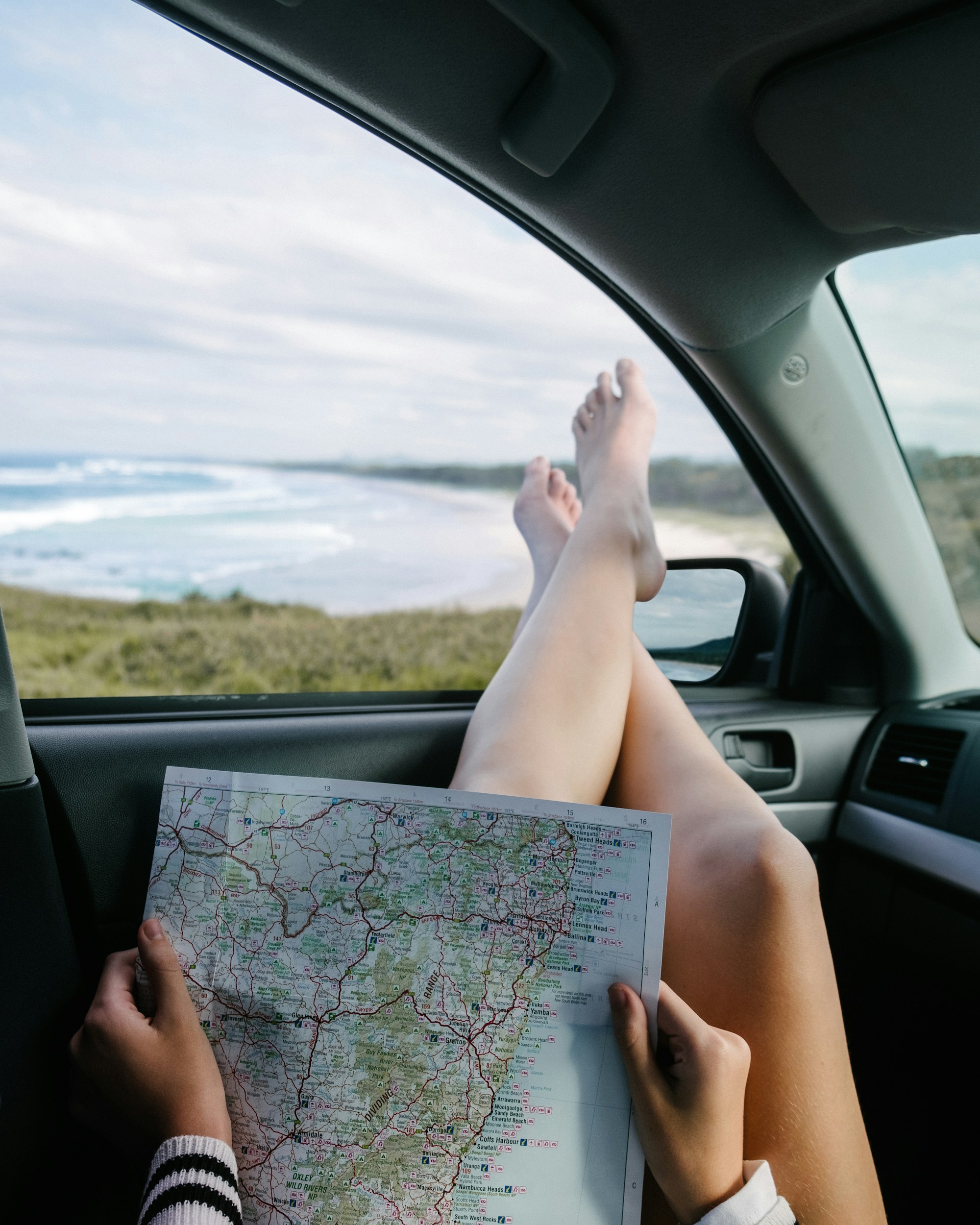 A passenger's bare feet up on the dashboard while their hands hold a paper road map, a beach and grassy headland outside