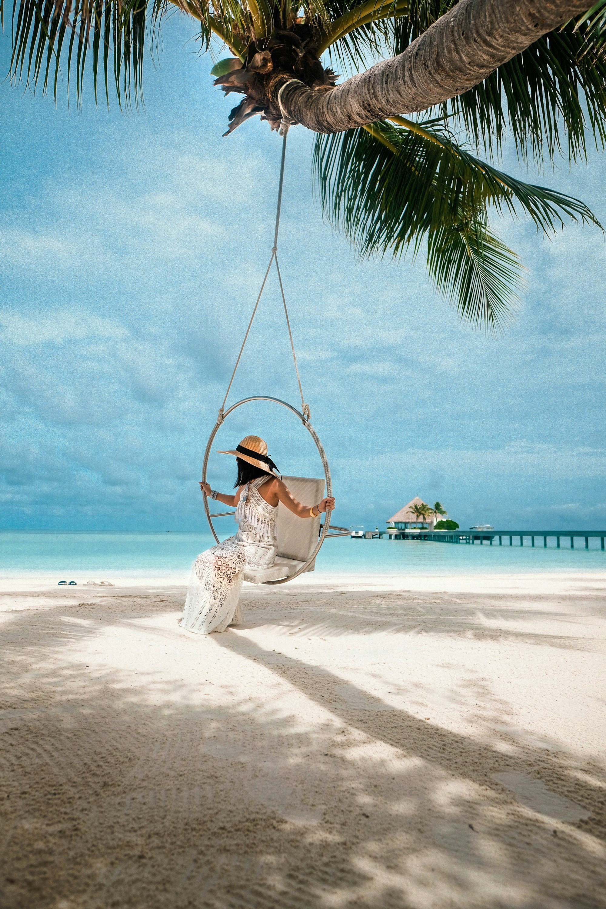 A woman in a long white crochet dress and a straw hat sitting on a hoop swing hanging from a palm tree, white sand and an overwater jetty behind