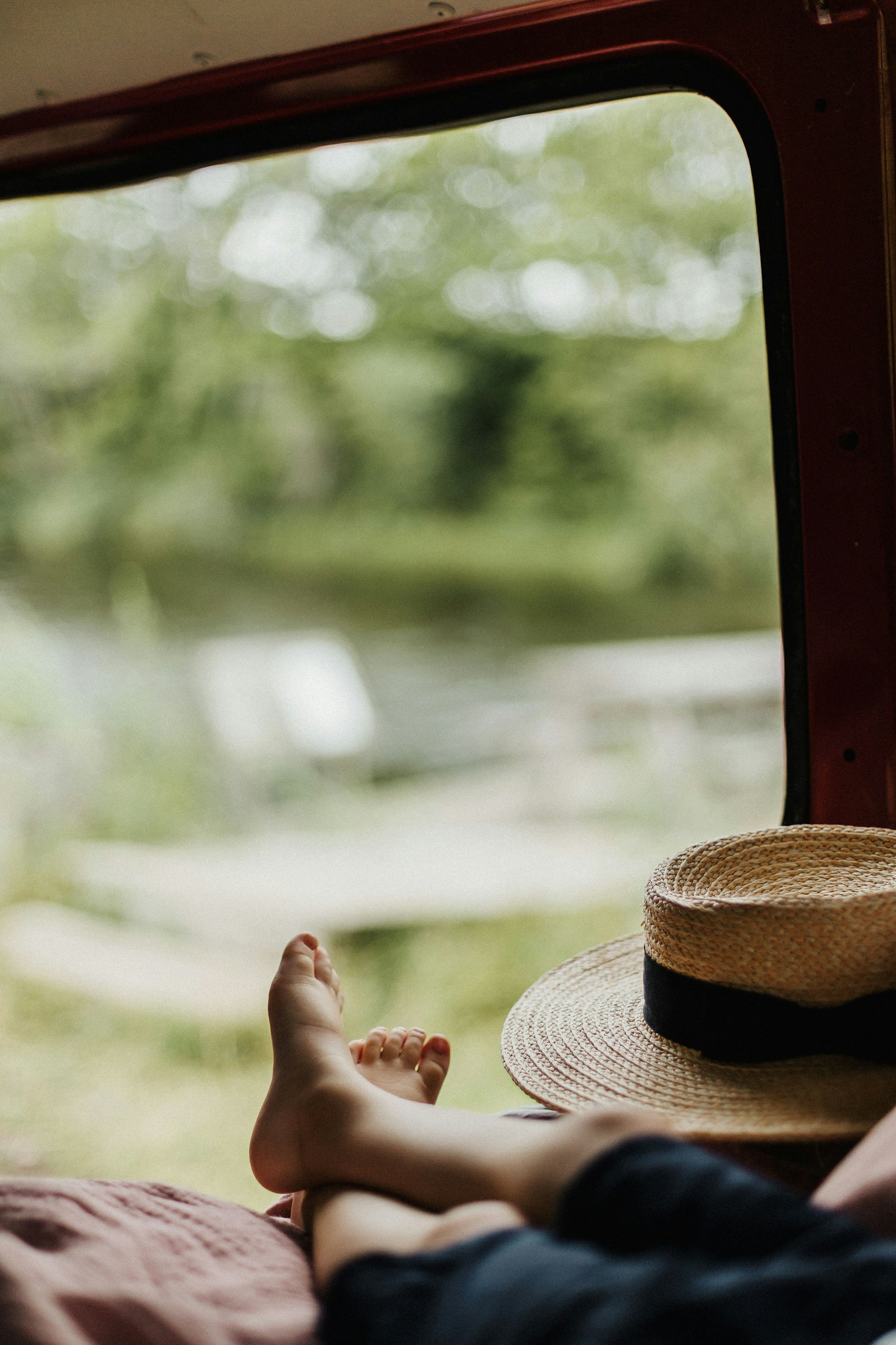 Bare feet and a straw boater hat inside a red-painted van parked beside green water and trees